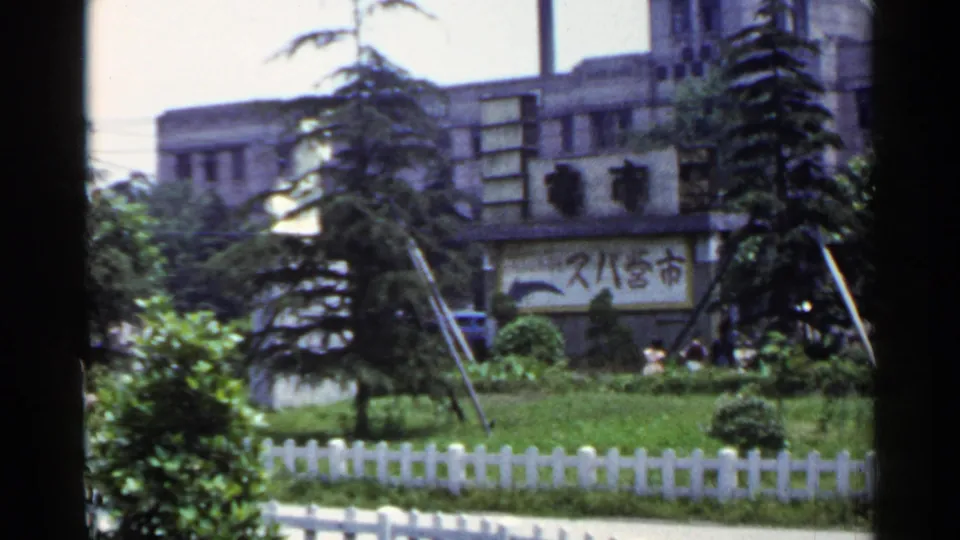 1953 Japan 8mm frame showing dense park greenery, a white fence, and a background urban building with Japanese characters visible.