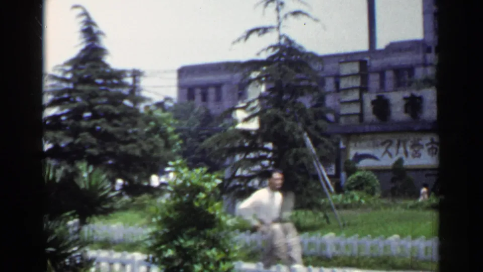 1953 Japan 8mm frame with a man in light traditional-looking clothing passing a fenced urban park beneath trees and Japanese signage.