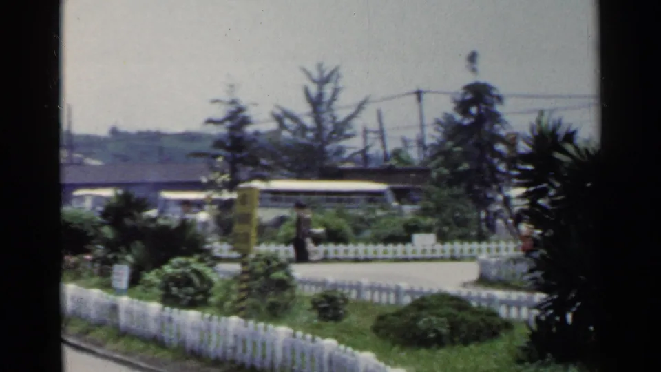 1953 Japan home-movie frame showing a white fence, garden shrubs, overhead wires and a covered roadside stop beyond the path.
