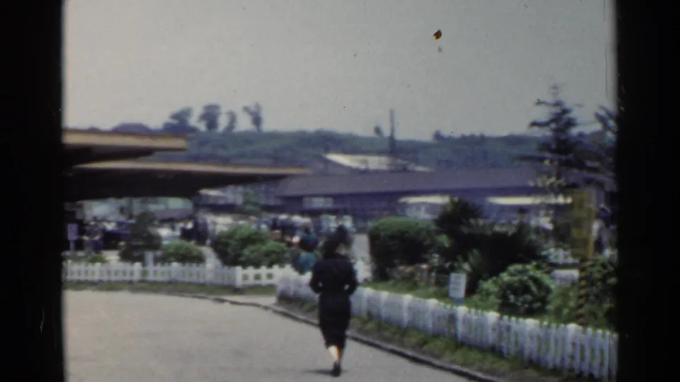 1953 Japan frame with a woman walking away on a paved path beside a white fence, shrubs and a covered roadside stop.