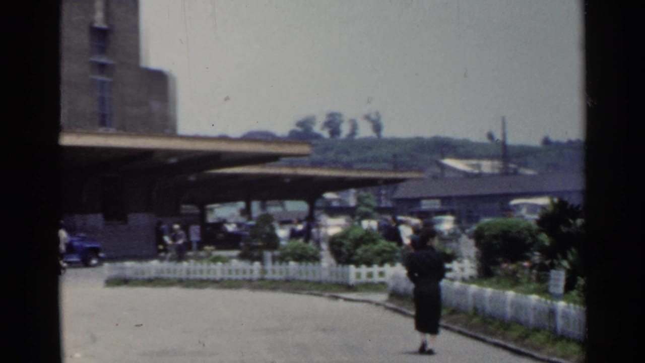 1953 Japan home-movie frame showing a brick terminal forecourt with buses, cars, shrubs, signs, and a woman walking by a white fence.