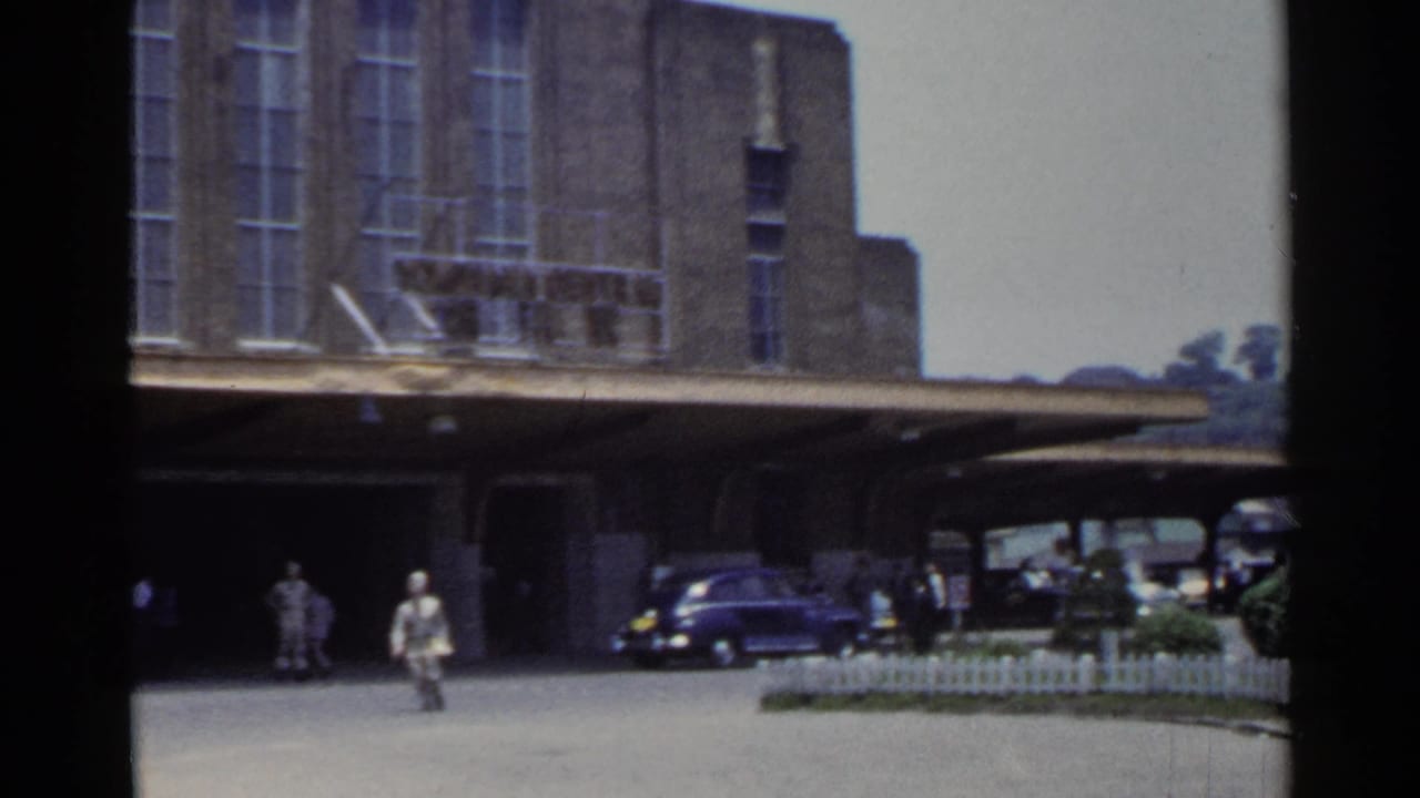 1953 Japan 8mm frame with pedestrians moving across a paved station forecourt beneath a large brick building and canopy.