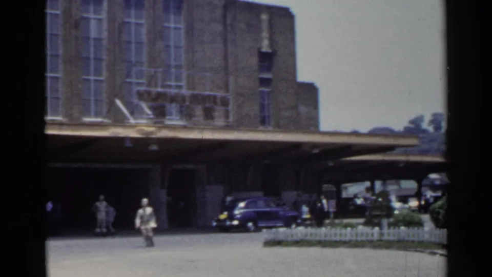1953 Japan 8mm frame with pedestrians moving across a paved station forecourt beneath a large brick building and canopy.