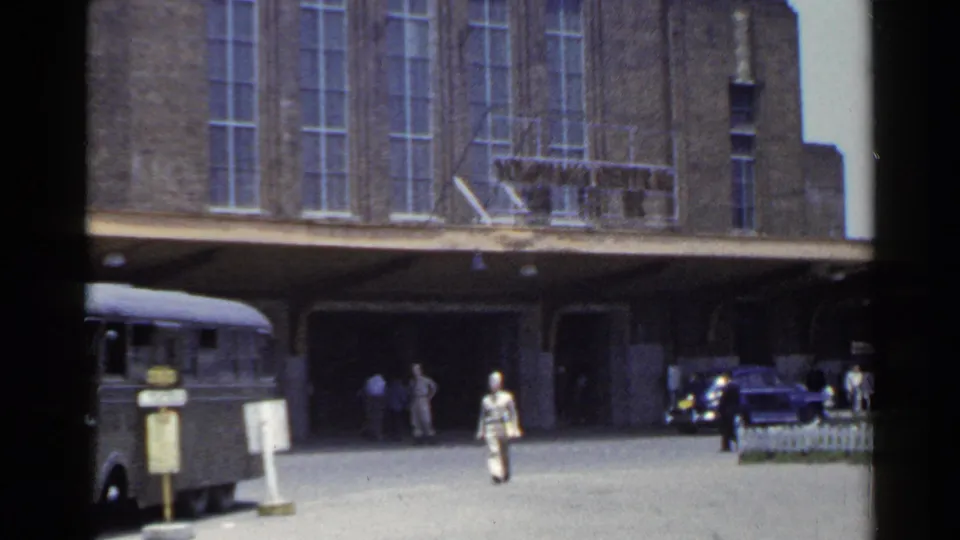 1953 Japan home-movie frame showing a bus at the curb, a light-suited passenger crossing, and cars under a brick terminal canopy.