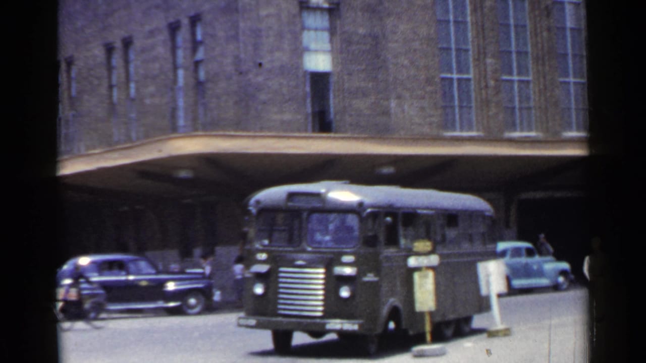 1953 Japan street view with a dark bus at a covered stop, period cars nearby, and a large brick building in the background.