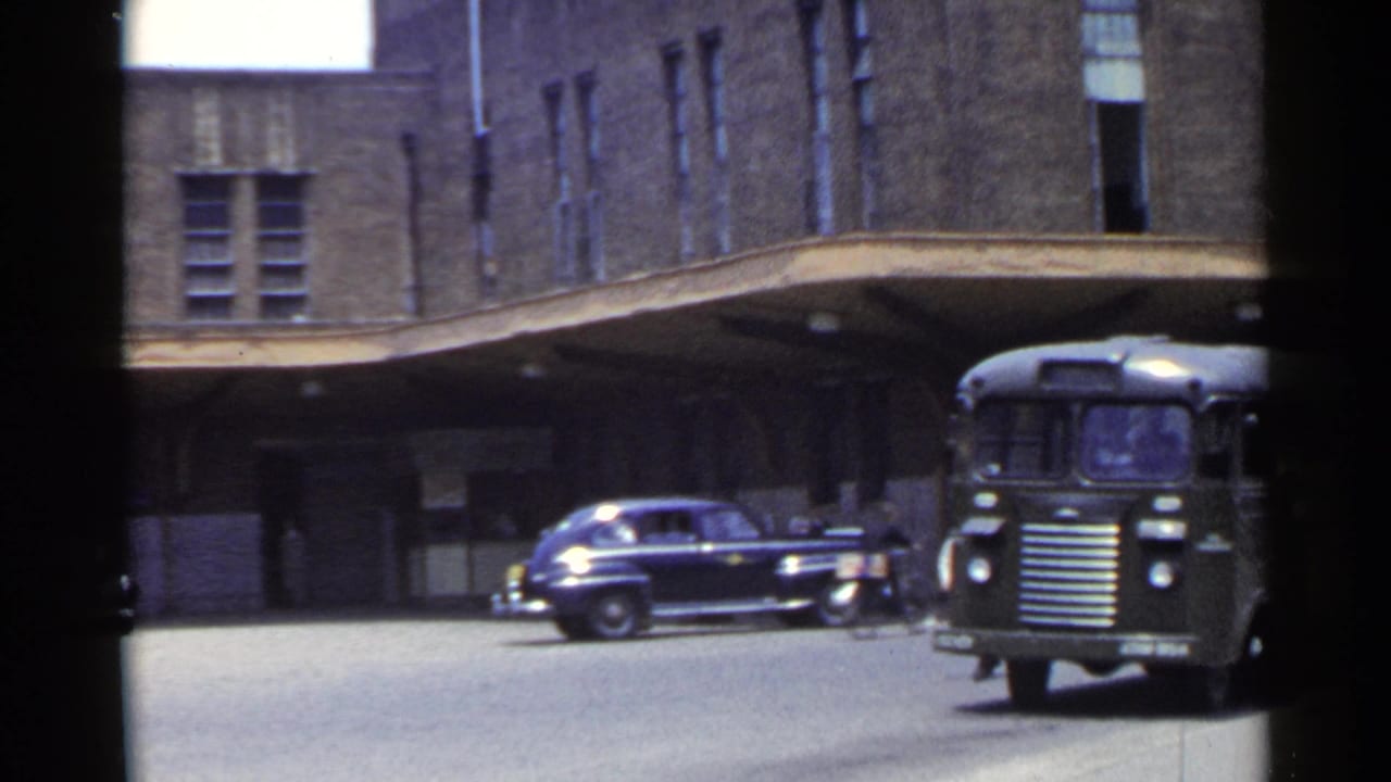 1953 Japan urban street frame showing a dark bus beside a covered building entrance and a period sedan crossing the scene.