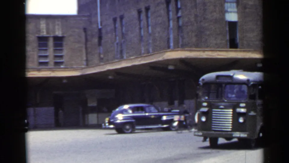 1953 Japan urban street frame showing a dark bus beside a covered building entrance and a period sedan crossing the scene.
