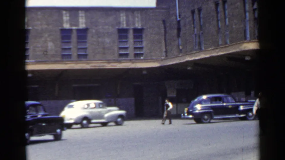 1953 Japan street scene with parked mid-century cars, a pedestrian crossing, and the covered base of a large brick building.