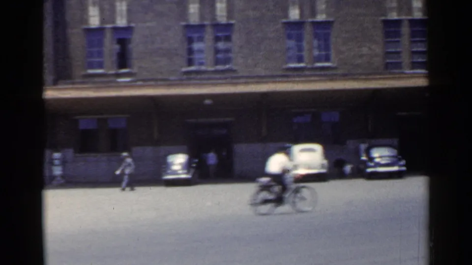 1953 Japan street frame with a cyclist passing through the foreground, pedestrians near the curb, and cars by a brick building.