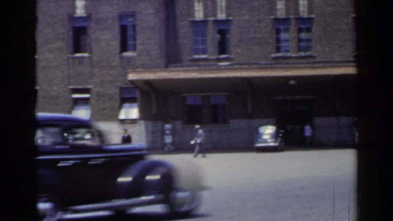 Dark sedan passing a brick building with pedestrians in a sunlit lot.
