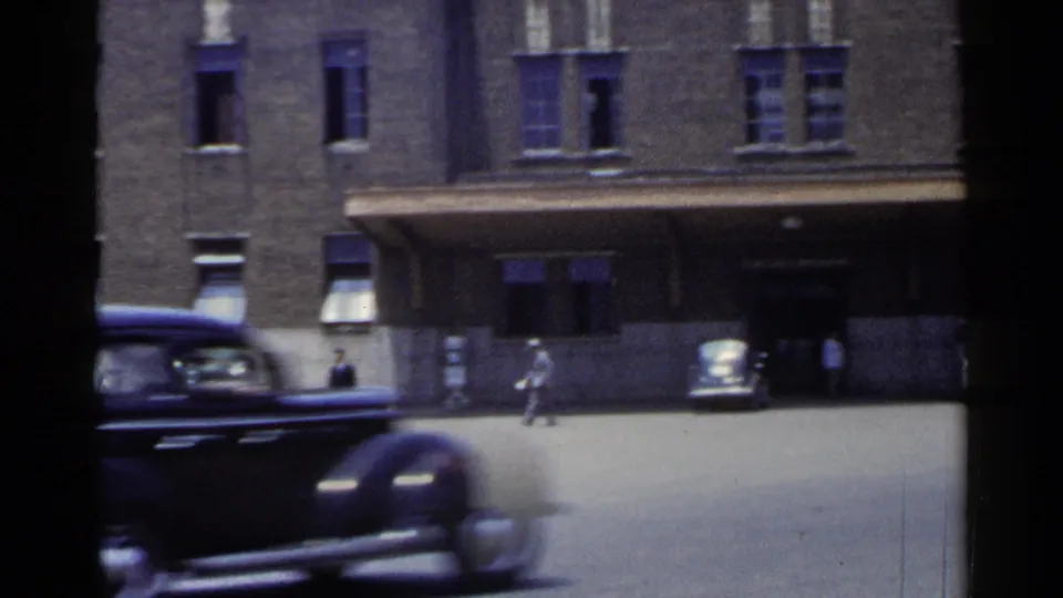 1953 Japan home-movie frame showing a dark sedan in front of a brick building as pedestrians cross a bright paved area.