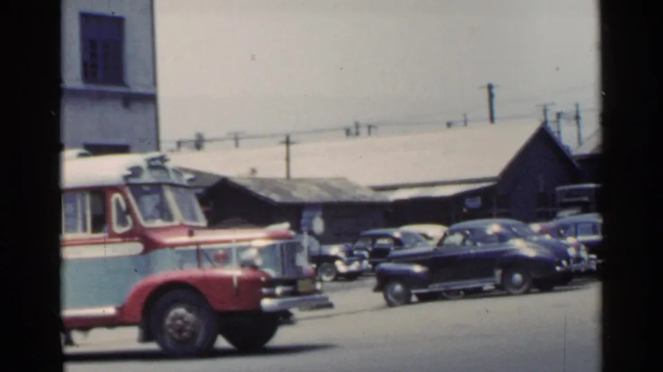 1953 Japan home-movie frame showing the front of a red-and-blue bus beside parked cars and low commercial buildings.