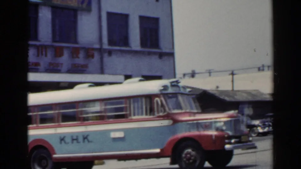 1953 Japan frame with a red-and-blue K.H.K. bus seen broadside below Japanese signage and windows.