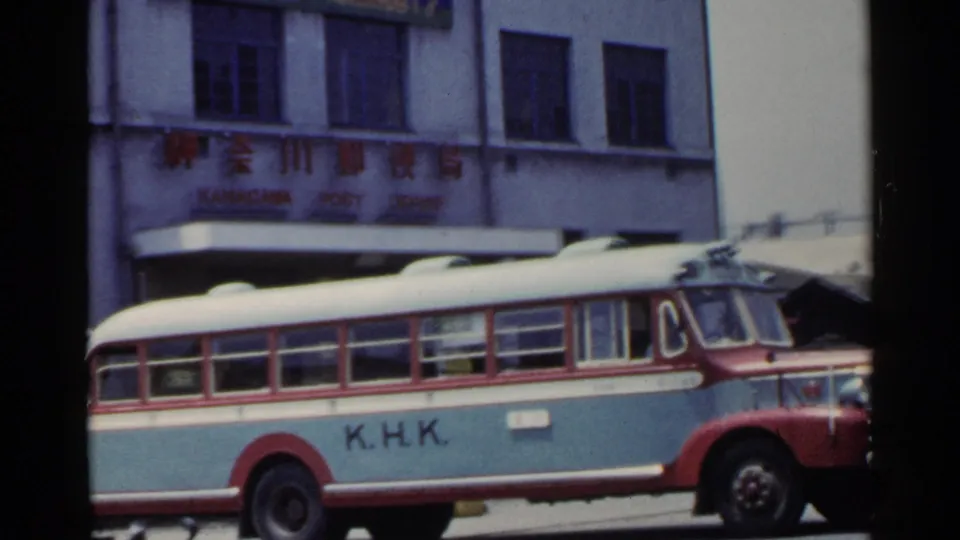 1953 Japan street view with a red, white, and light blue K.H.K. bus passing a building marked with Japanese signage.