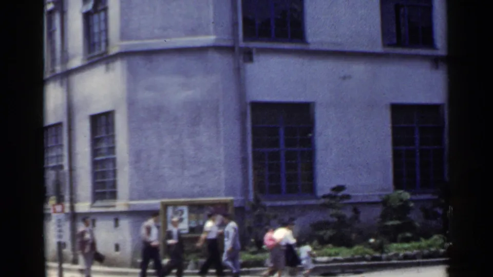 1953 Japan sidewalk view with several pedestrians outside a large building with tall windows and planted edging.