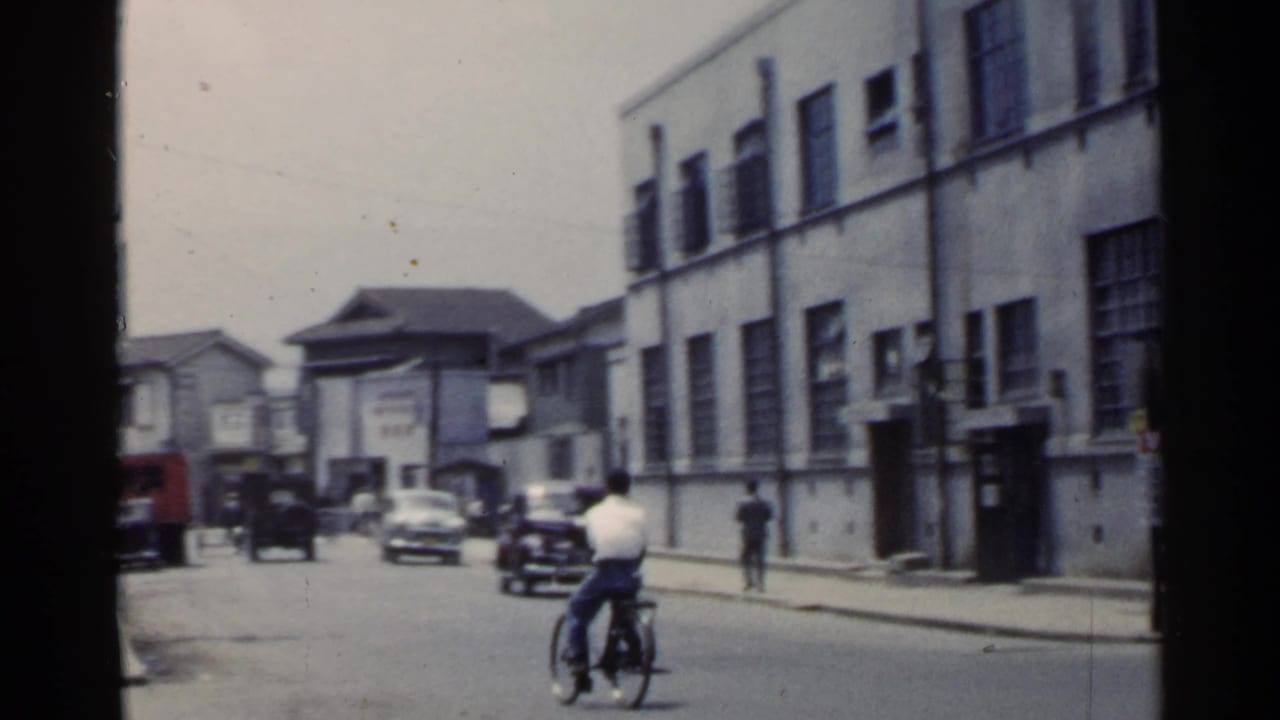 Cyclist rides down a town street past cars, pedestrians, and low commercial buildings.