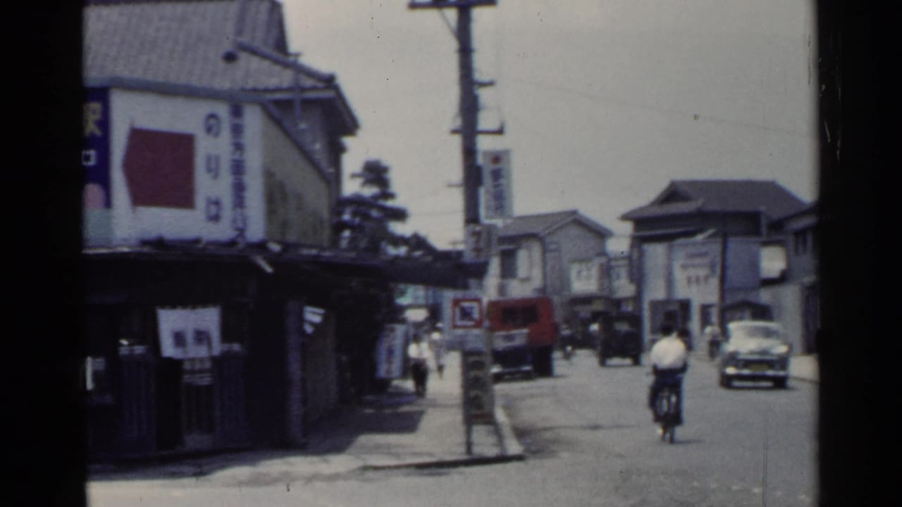 1953 Japan street corner showing a bicyclist, cars, pedestrians, Japanese signage, and overhead utility poles.