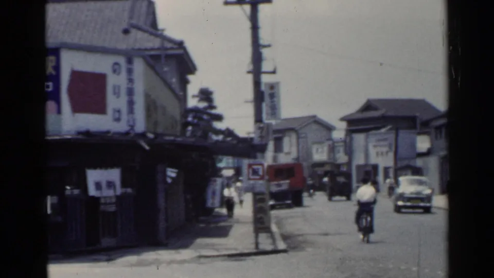 1953 Japan street corner showing a bicyclist, cars, pedestrians, Japanese signage, and overhead utility poles.