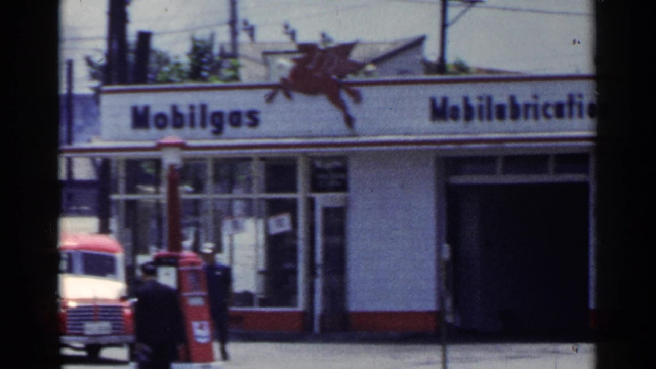 1953 Japan 8mm frame showing a Mobilgas service station facade with Pegasus branding, service bay, pedestrian, and red vehicle near the pumps.