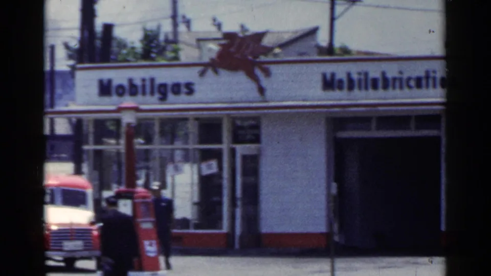 1953 Japan 8mm frame showing a Mobilgas service station facade with Pegasus branding, service bay, pedestrian, and red vehicle near the pumps.