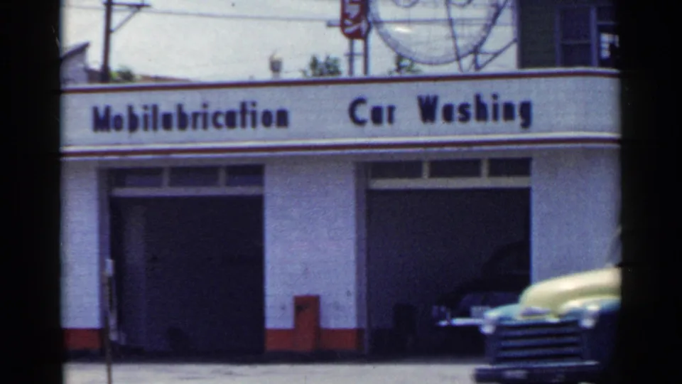 1953 Japan 8mm frame showing Mobilubrication and car washing signage with a light-colored mid-century vehicle passing in front.