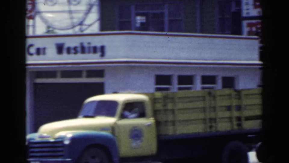 1953 Japan 8mm frame showing a yellow delivery truck moving past the Mobilgas car washing area and service station frontage.