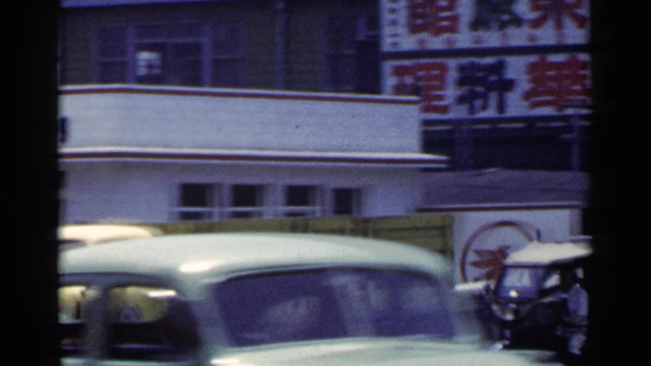 Classic car passing in front of storefront signs and a small vehicle.
