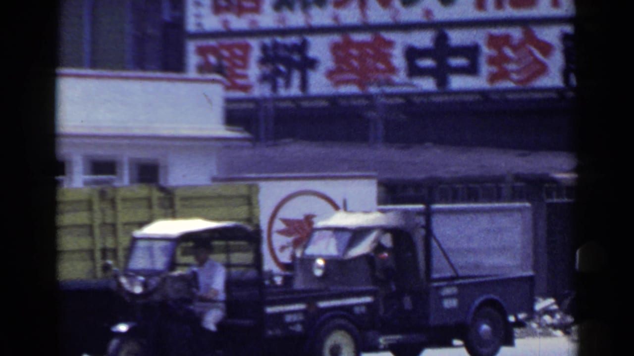 1953 Japan street frame showing a dark delivery truck, a smaller vehicle, a pedestrian, and large Chinese-character signs above the storefront.