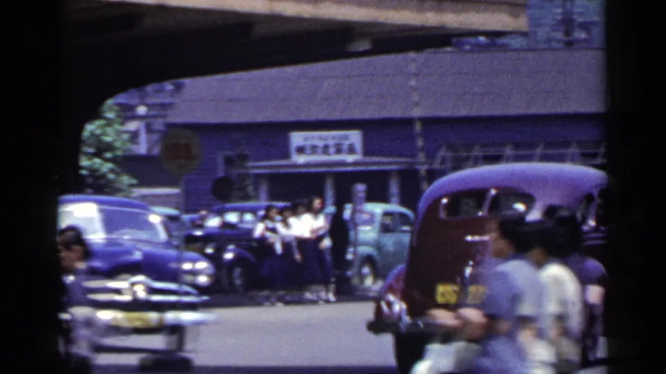 1953 Japan street scene with pedestrians by parked classic cars, including a purple car and a reddish vehicle in the foreground.