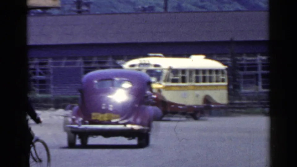 1953 Japan street frame with a purple classic car in the roadway, a cream-and-yellow bus behind it, and a building facade in the background.