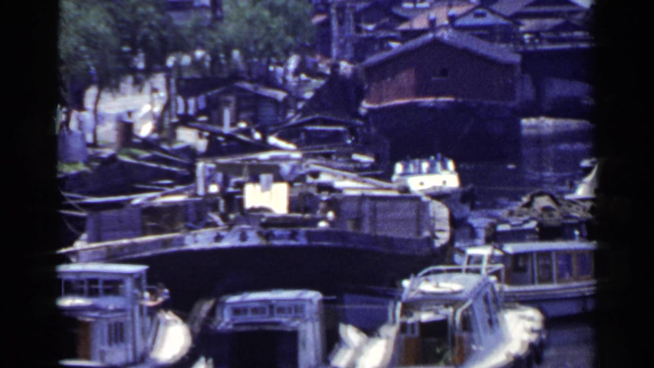 1953 Japan 8mm frame showing a dense canal scene with moored boats, wooden waterfront structures, and a bridge beyond.
