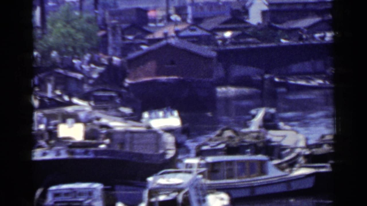Small boats and houseboat-like cabins crowd a canal near wooden structures.