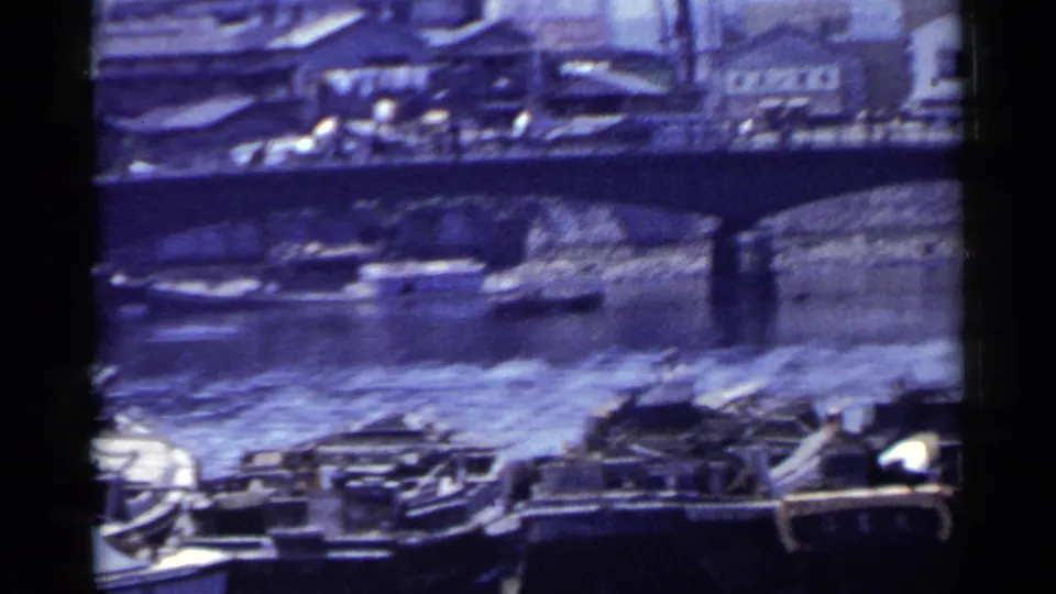 1953 Japan home-movie frame showing a canal bridge, moored boats, water reflections, and dense waterfront structures.