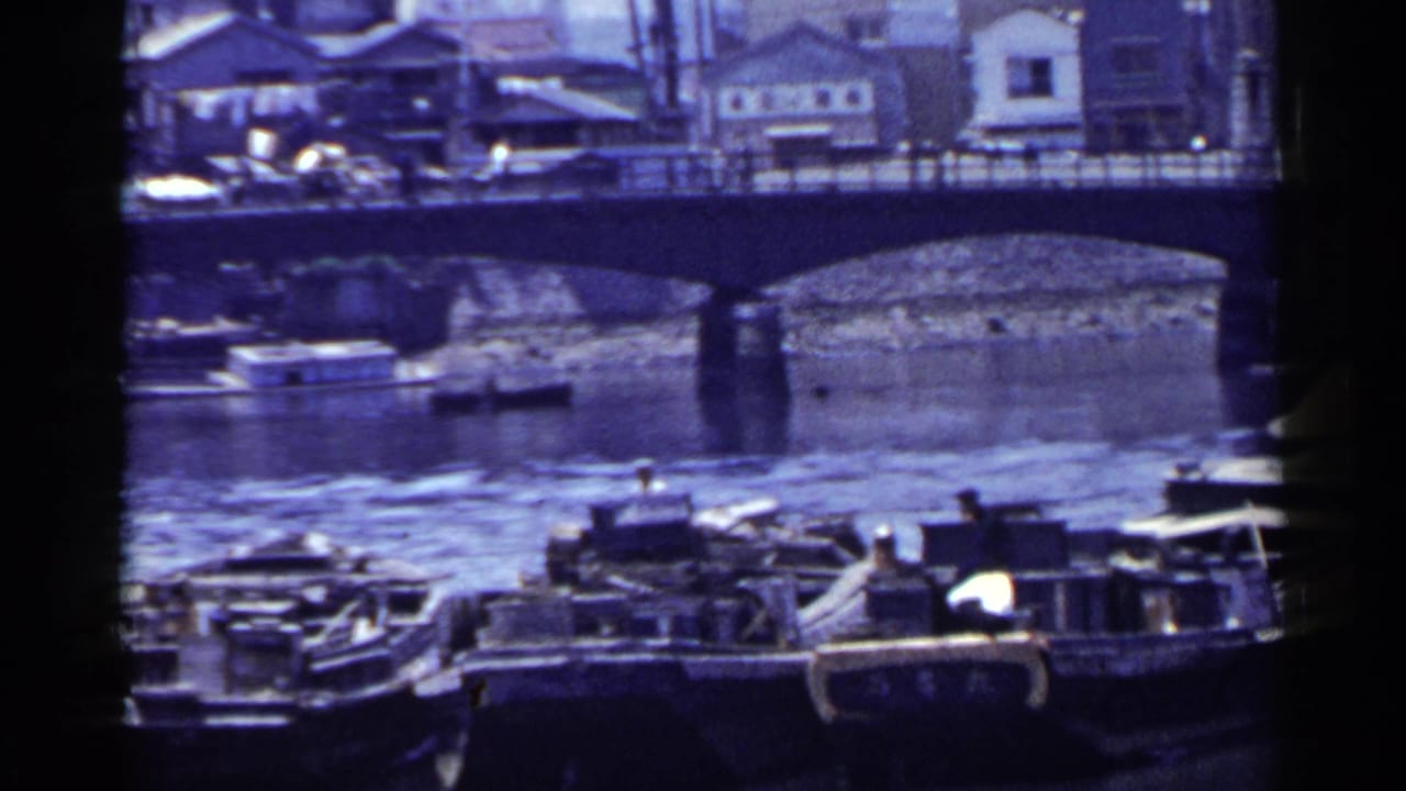 1953 Japan home-movie frame showing small boats on a river beneath an arched bridge with houses along the opposite bank.