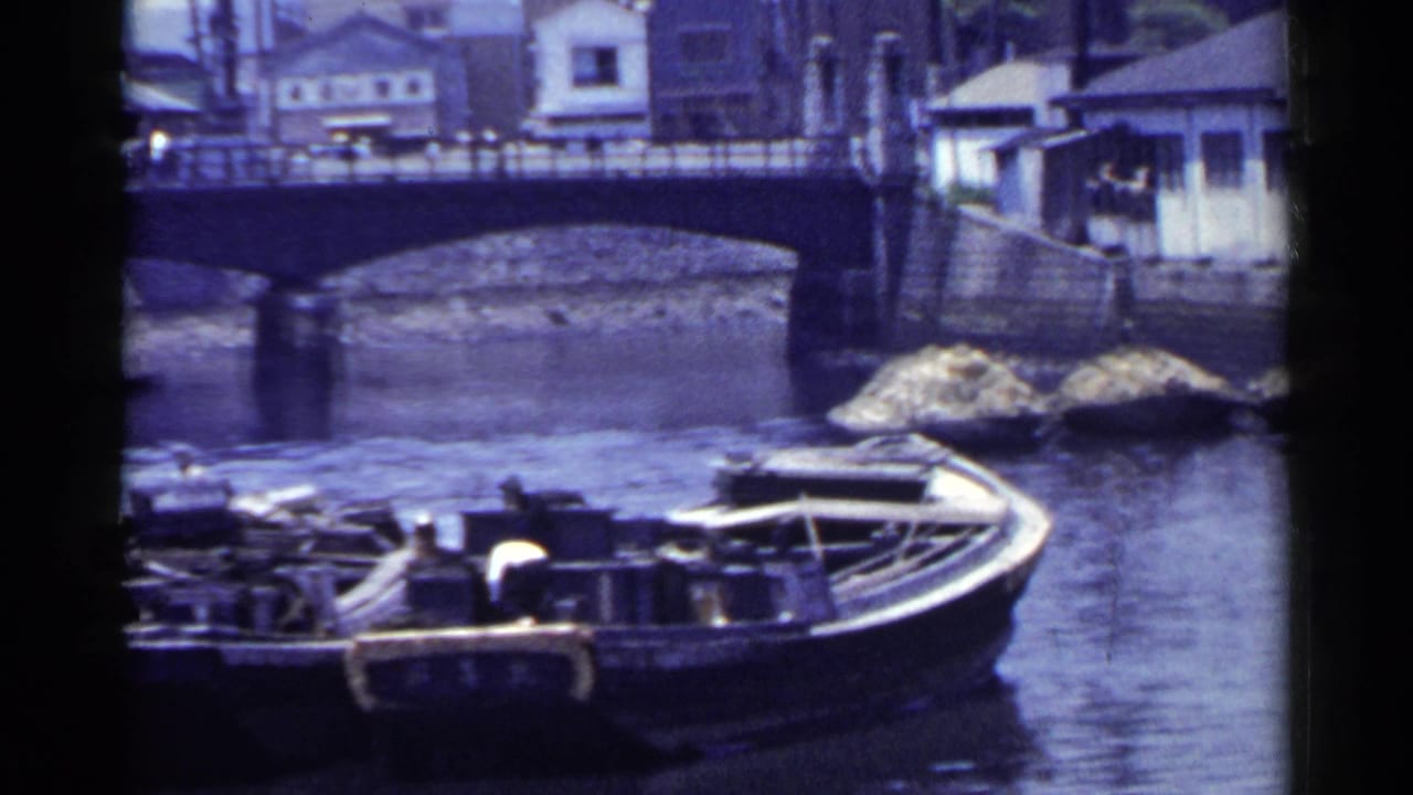 1953 Japan river scene with a large moored boat in the foreground, an arched bridge, stone bank, and waterside buildings behind.
