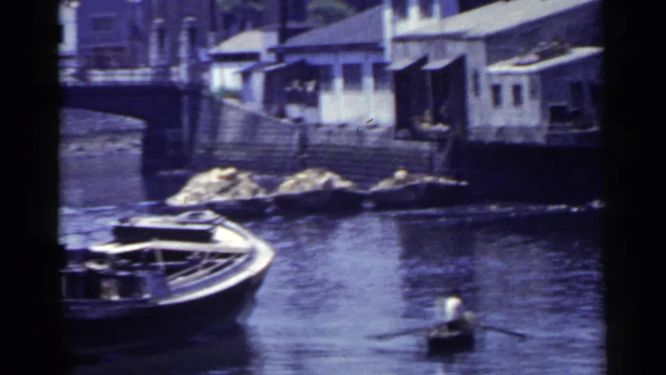 1953 Japan home-movie view of a boat along the river edge, with waterfront houses, steps, rocks, and a partial bridge at left.