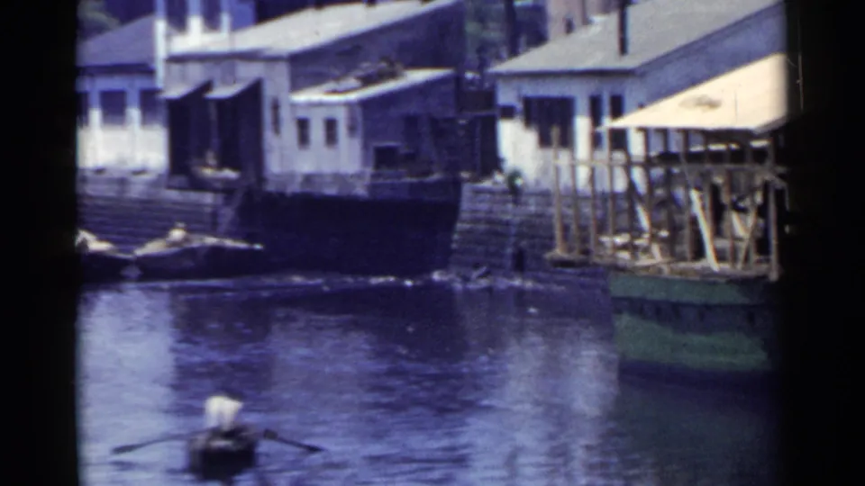 1953 Japan frame showing a small rowboat on the river beside waterfront buildings, rocks, and a raised dock or pier structure.
