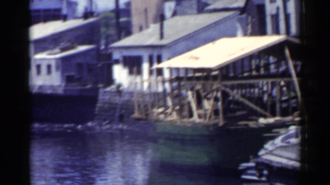 1953 Japan home-movie frame showing a covered wooden dock over calm water with industrial waterfront buildings behind it.