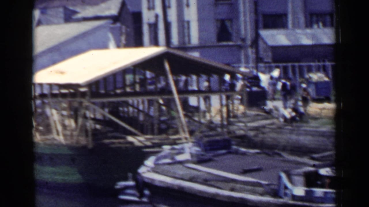 1953 Japan waterfront frame with a covered dock, stone edge, pedestrians, and stacked cargo or material near industrial buildings.