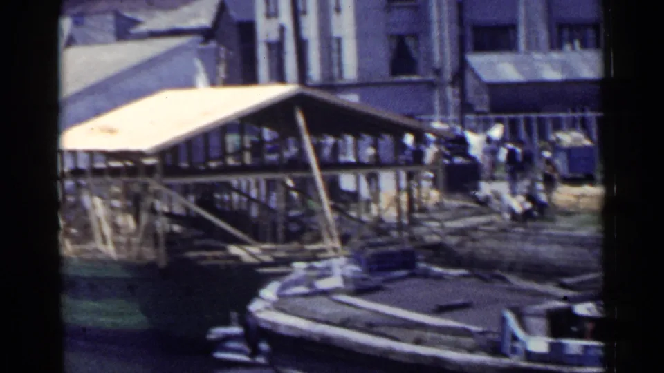 1953 Japan waterfront frame with a covered dock, stone edge, pedestrians, and stacked cargo or material near industrial buildings.