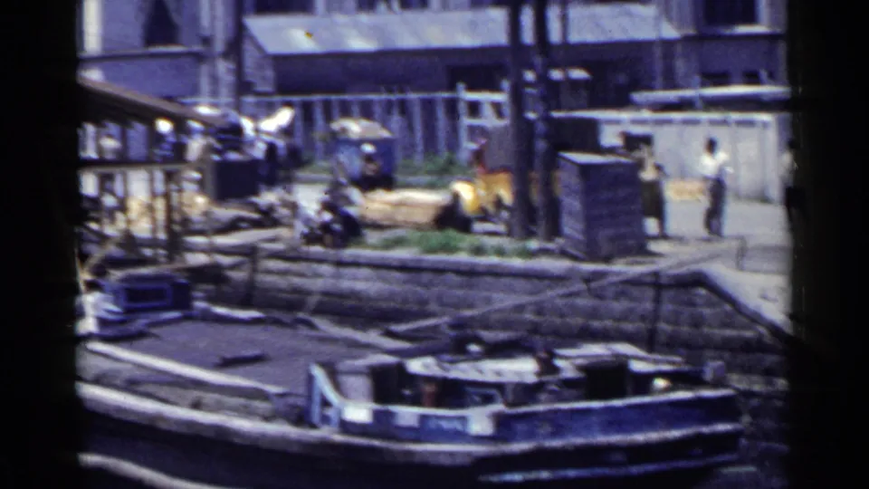 1953 Japan home-movie frame showing a moored boat, stone steps, pedestrians, a yellow truck, and dockside cargo.