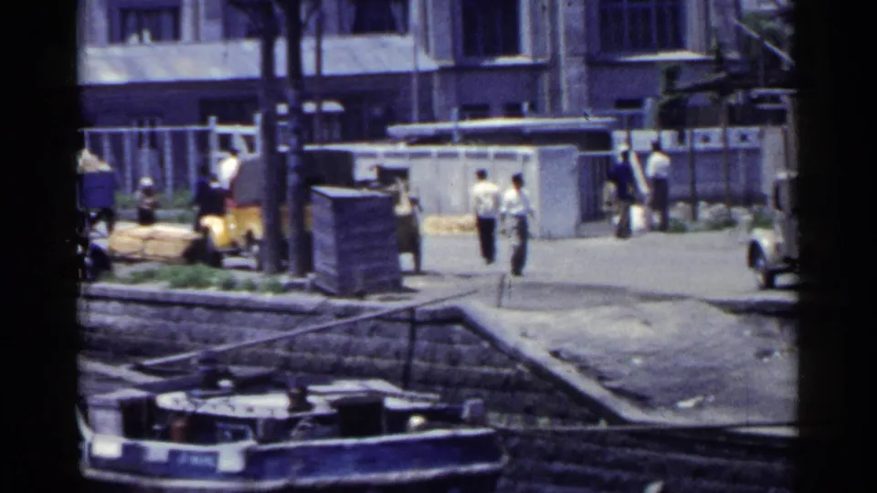 1953 Japan waterfront frame with pedestrians on the dockside street, a moored boat below, and industrial buildings in the background.