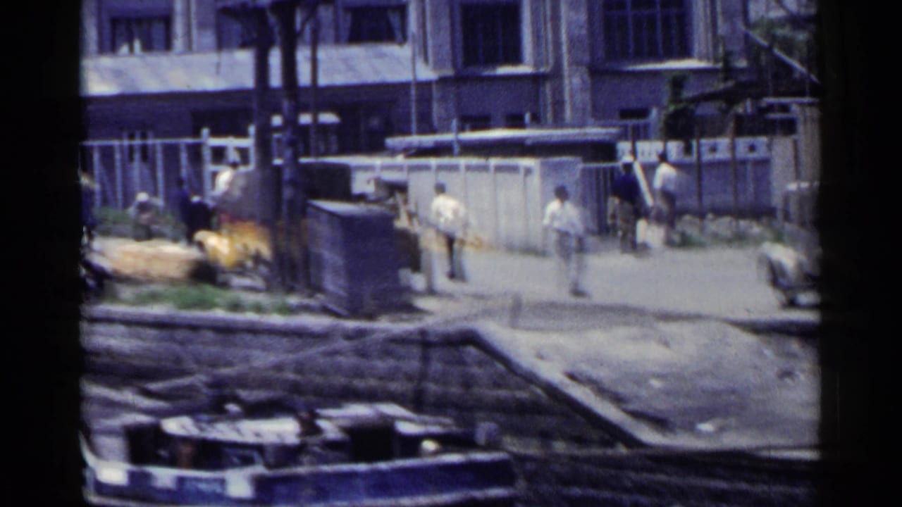 Urban street scene with pedestrians, buildings, a bridge railing, and a yellow vehicle in the distance.