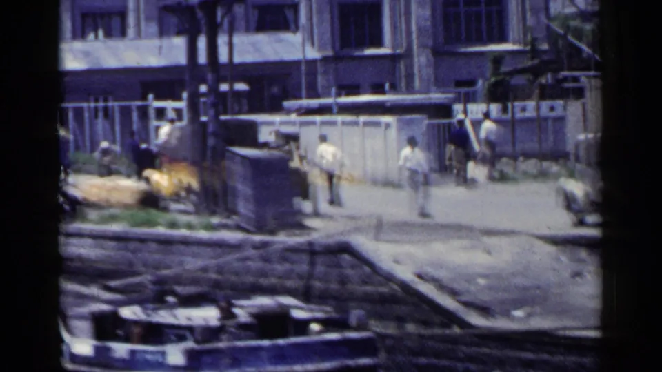 1953 Japan 8mm street view with pedestrians near buildings, a stone railing, and a yellow vehicle in a busy urban setting.