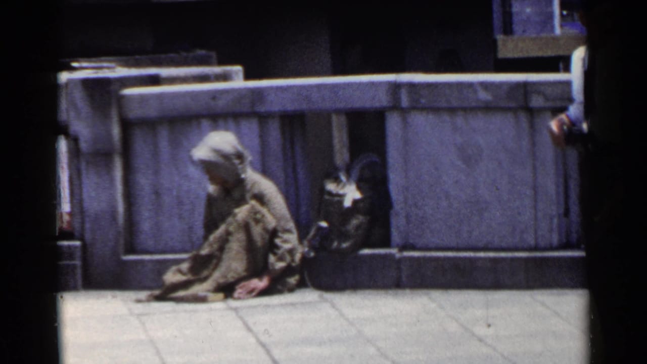 Seated man in worn clothing on a Japanese urban sidewalk in 1953, with a stone wall and dark bag nearby.