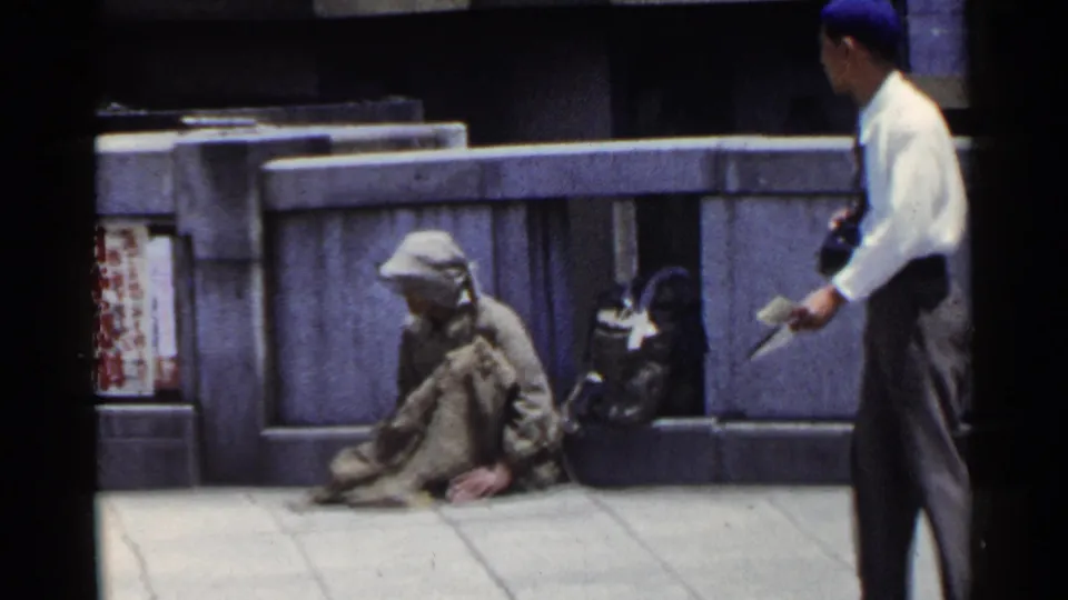 1953 Japan sidewalk view with a seated man against a stone wall while a pedestrian passes through the foreground.