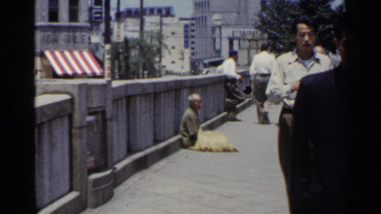 1953 Japan street footage shows pedestrians on a stone bridge walkway passing a seated person near the railing and striped awning.