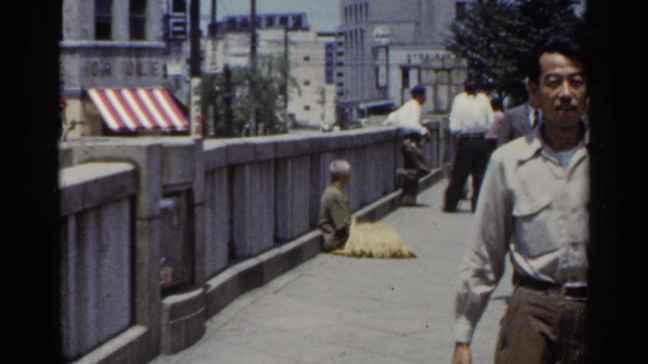 In a 1953 Japan street scene, a man in a light shirt approaches camera while a seated figure remains near the stone bridge railing.