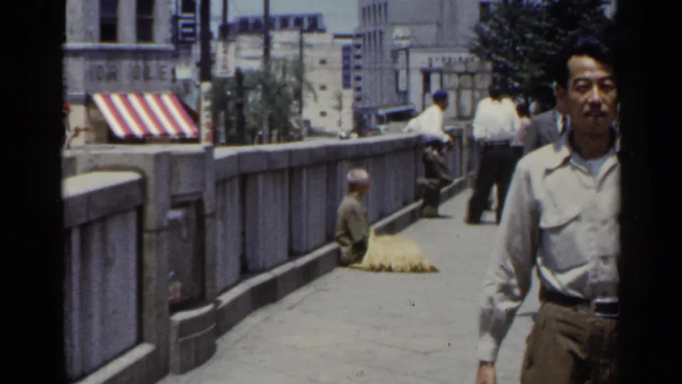 In a 1953 Japan street scene, a man in a light shirt approaches camera while a seated figure remains near the stone bridge railing.