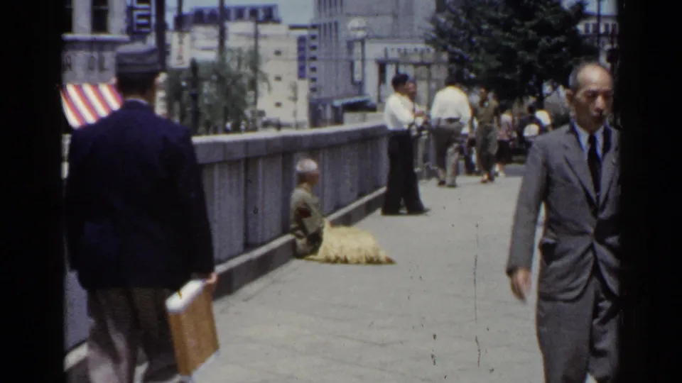 Pedestrians cross the frame in 1953 Japan footage beside a stone bridge railing, with a seated person and striped awning visible behind them.